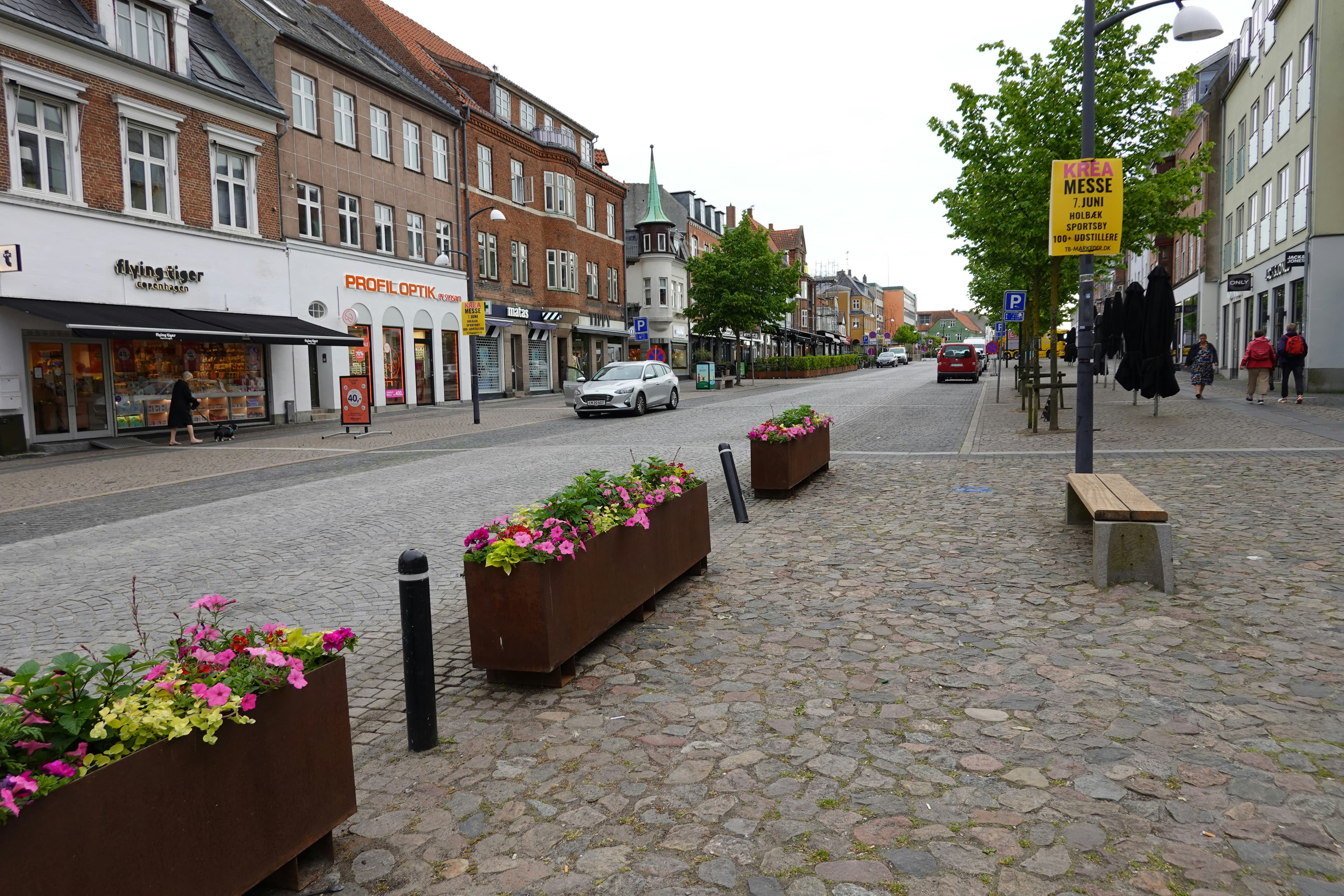 Brostensbelagt handelsgade med butiksfacader og blomsterkummer